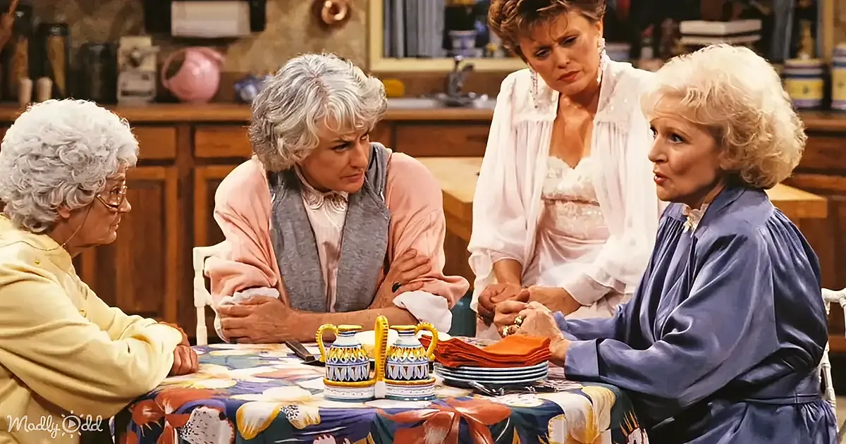 Four women sit around a table with a floral tablecloth, engaged in conversation in a cozy kitchen setting.