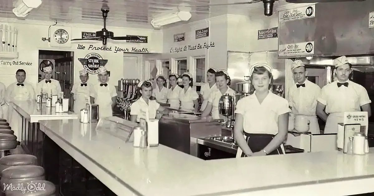 Waitresses in white uniforms stand behind a counter in a diner, with a sign reading "We Protect Your Health."