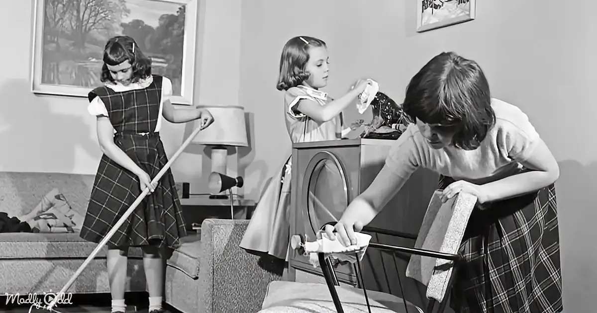 Three girls in plaid dresses clean a living room, one sweeps, another wipes, and the third polishes furniture.