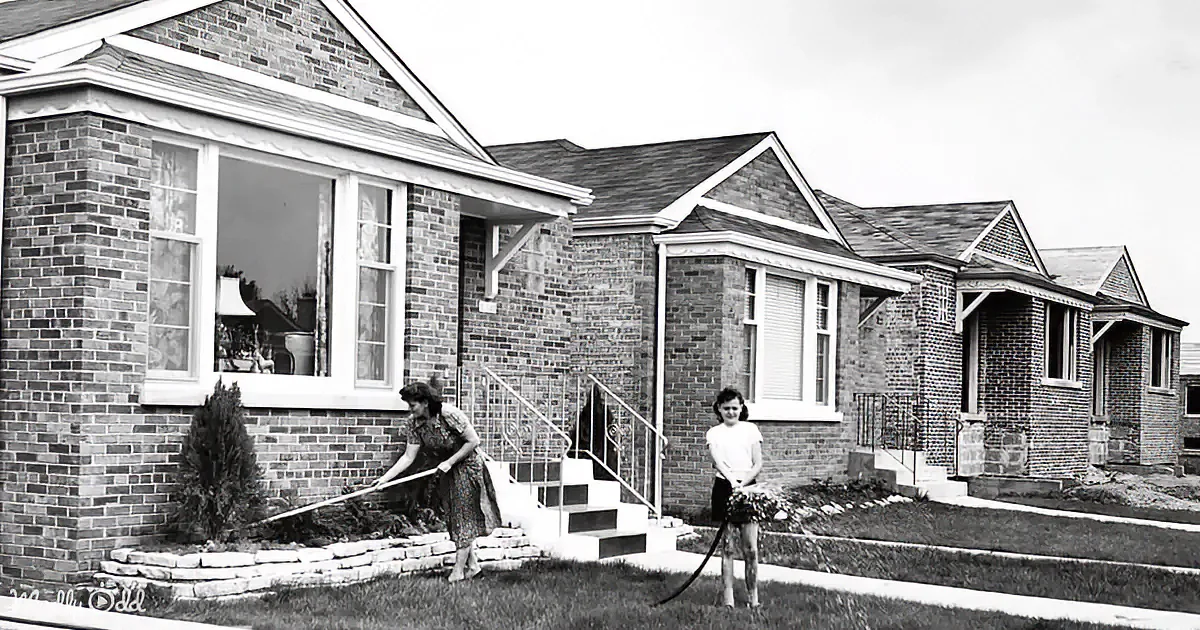 A woman and child water plants in front of a brick house with a row of similar homes in the background.