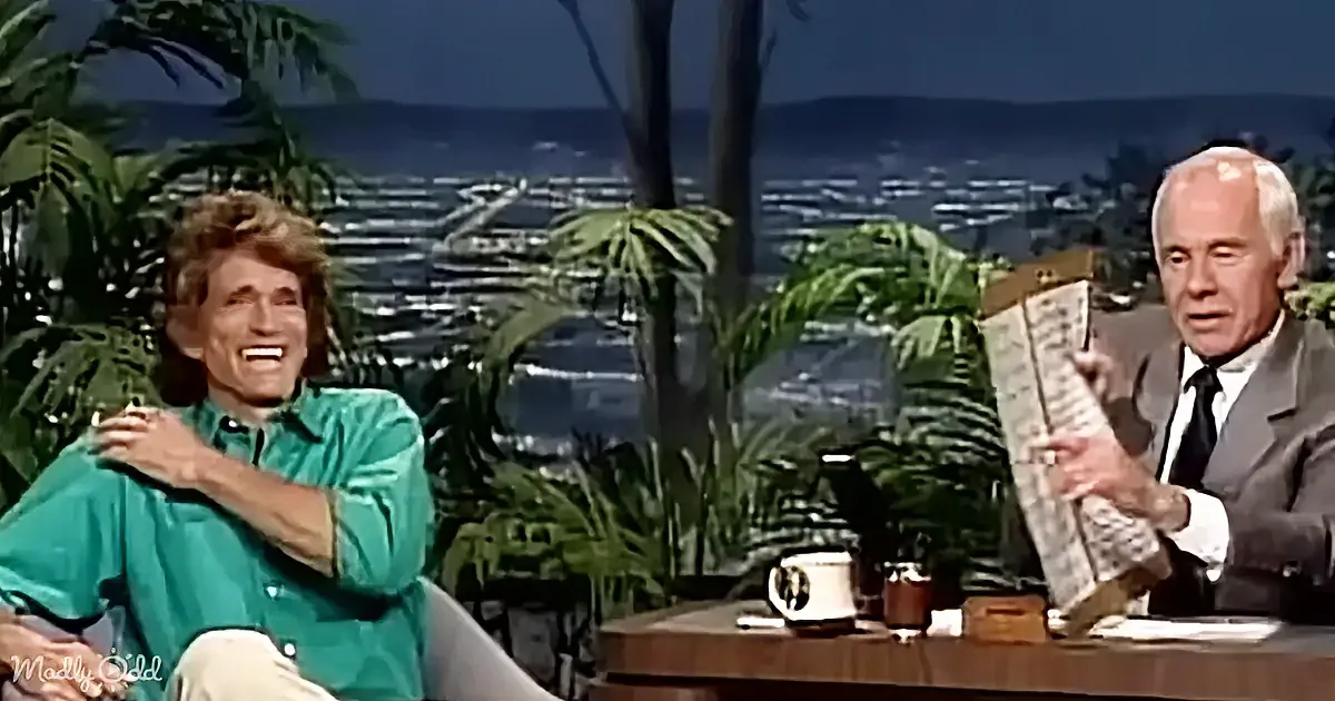 A man in a green shirt laughs while sitting next to a man in a suit holding a clipboard.