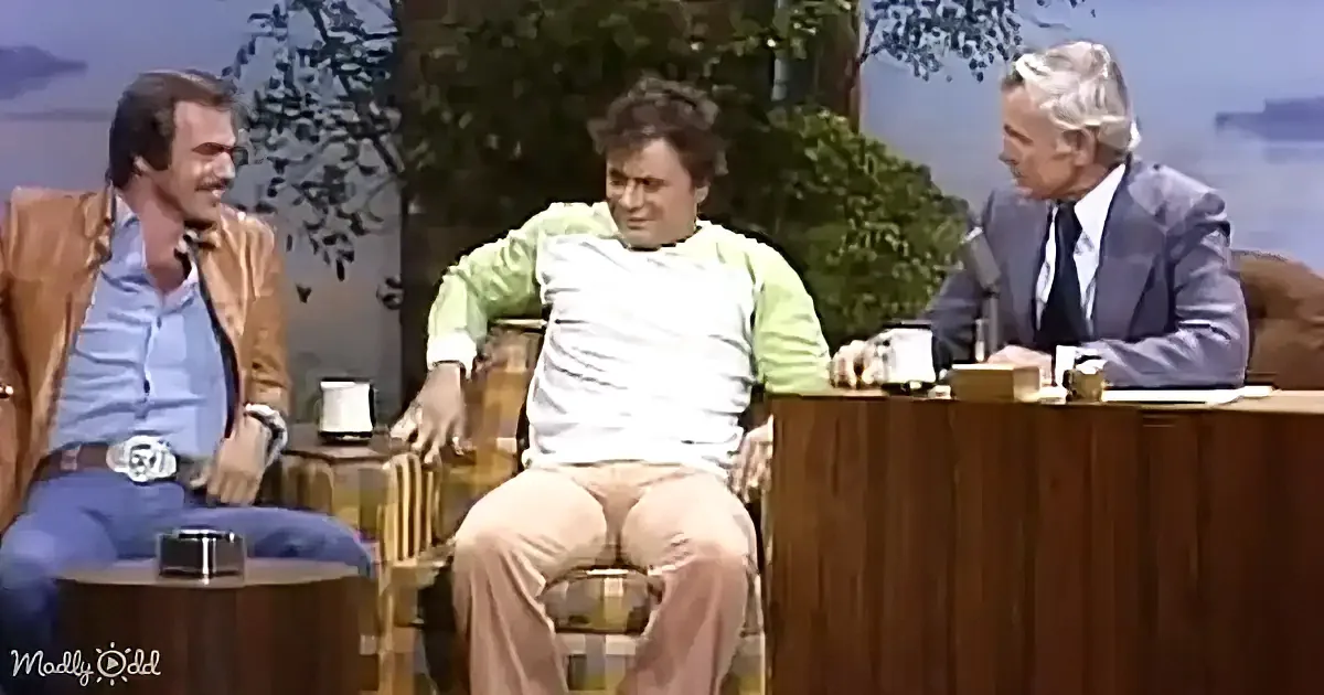 A man in a green shirt sits on a stool, talking to another man in a brown jacket and a man in a suit at a desk.