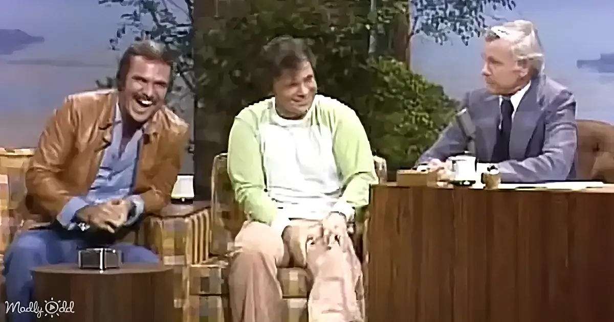 Three men are seated on a talk show set, with one in a brown jacket laughing, another in a green shirt, and a third in a...