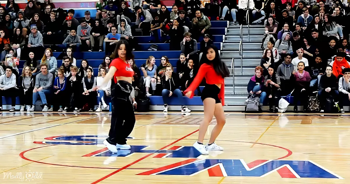 Two dancers in red and black perform on a basketball court with a crowd of spectators watching.