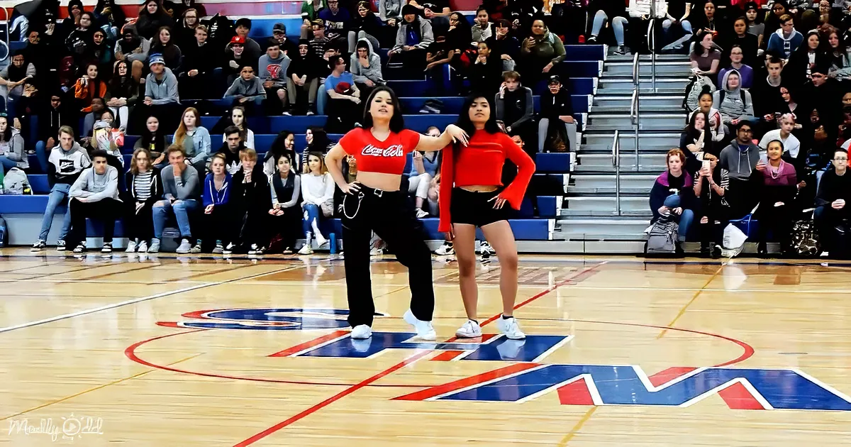 Two dancers in red tops and black pants pose on a basketball court with a crowd watching.