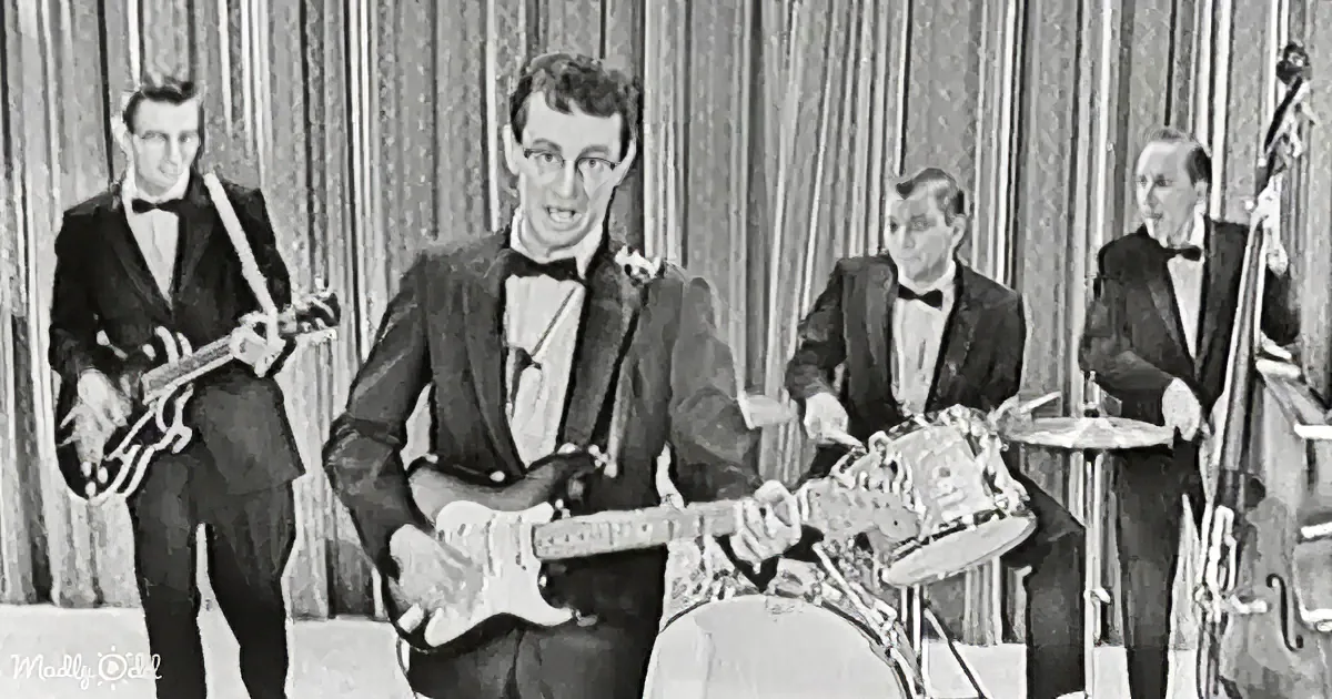 A man in a suit plays guitar while a band in tuxedos performs in front of a striped curtain.