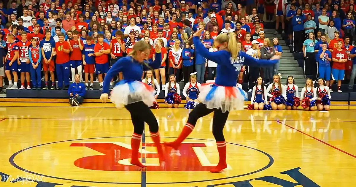 Two cheerleaders in blue and red outfits perform a routine on a basketball court with a crowd watching.