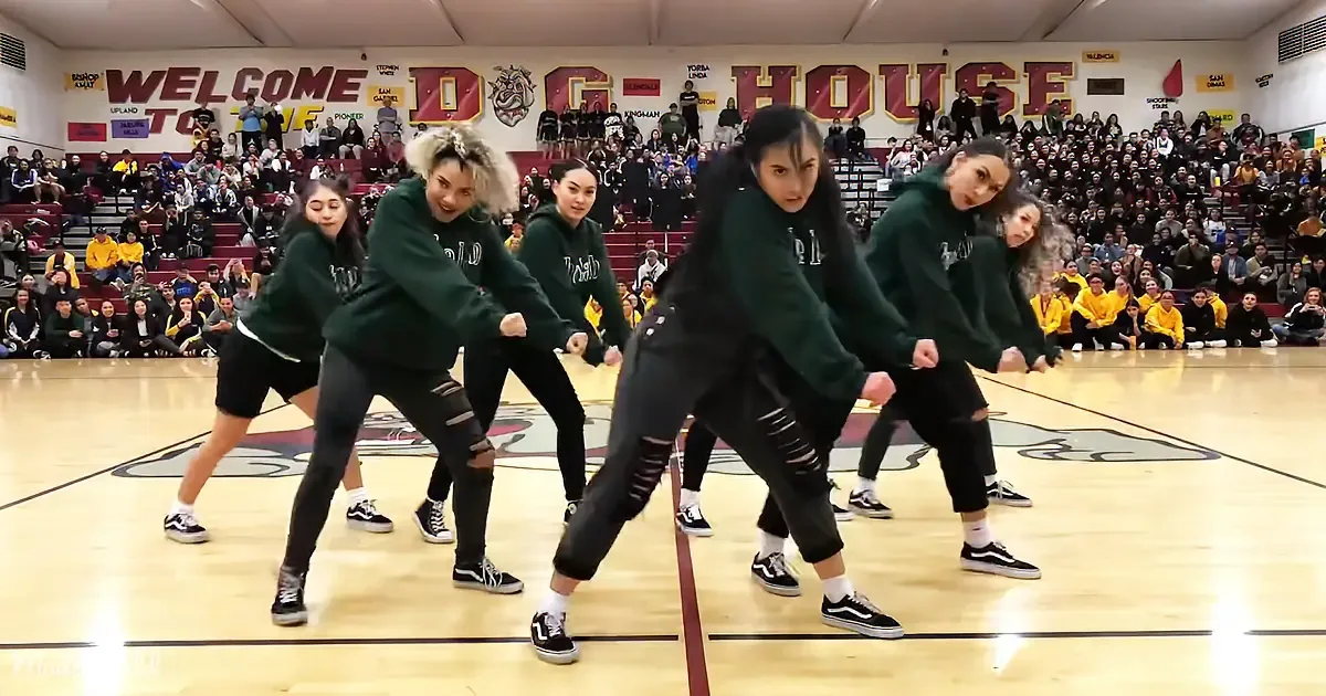 A group of dancers in green hoodies perform a synchronized routine on a gymnasium floor.