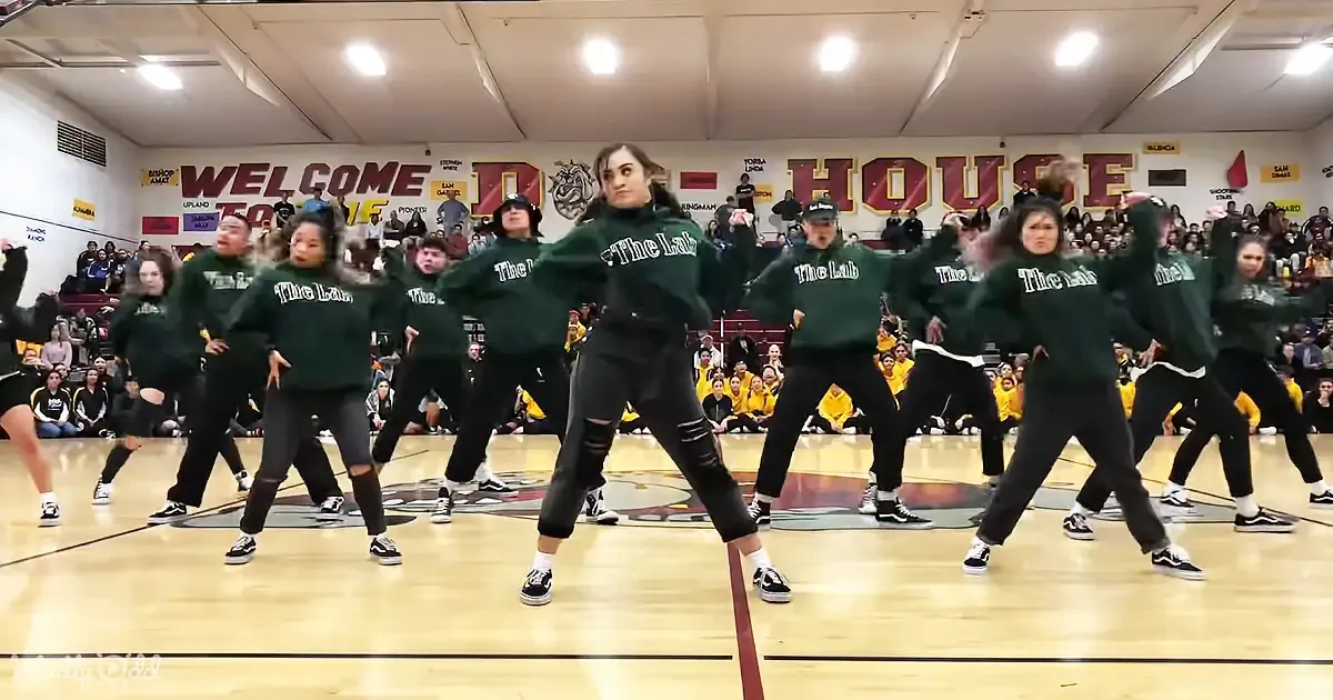 A group of dancers in green "The Lab" sweatshirts perform a synchronized routine on a gymnasium floor.