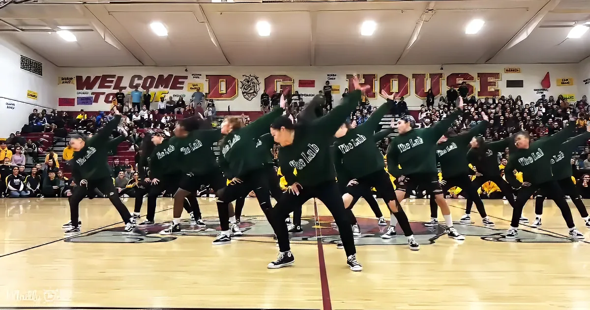 A synchronized dance group in green "The Lab" sweatshirts performs on a gymnasium floor, with spectators in the background.