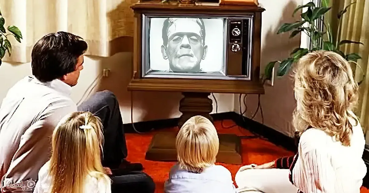 A family watches an old TV showing Frankenstein's monster, sitting on a red carpet.
