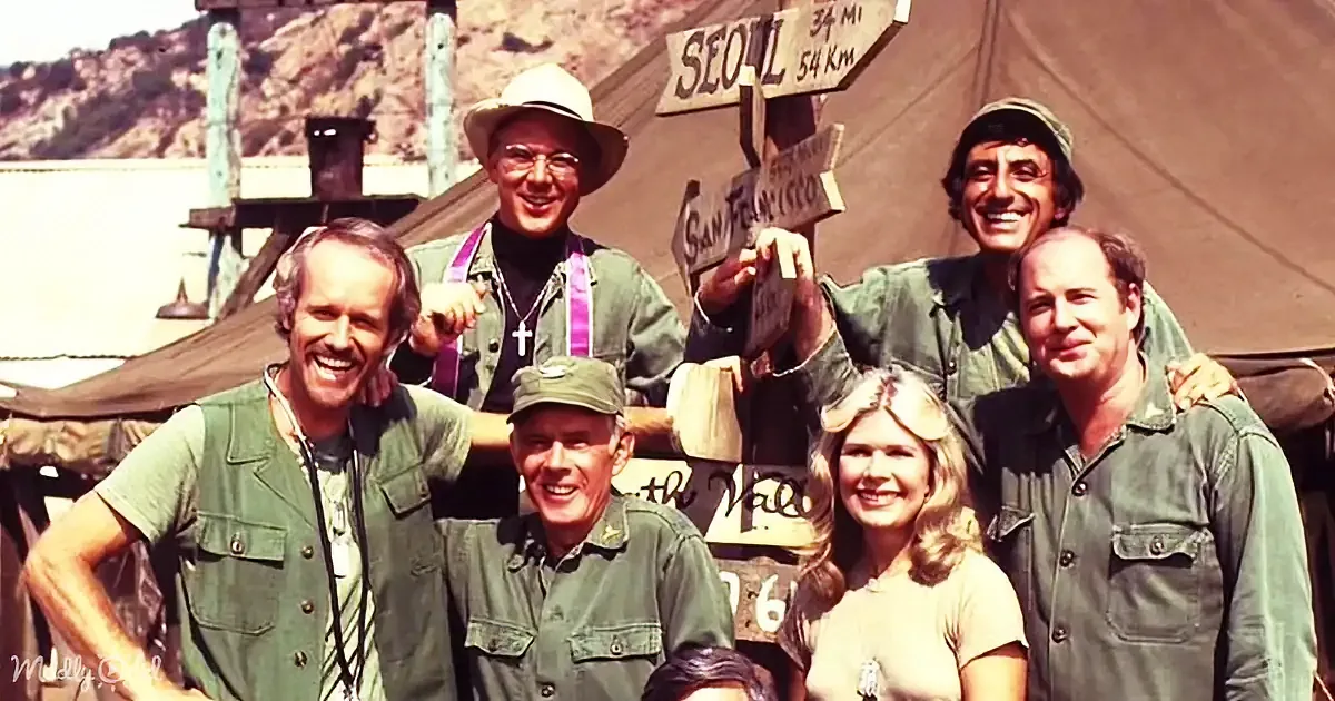 Six people in green attire smile near a signpost with distances to Seoul and San Francisco.