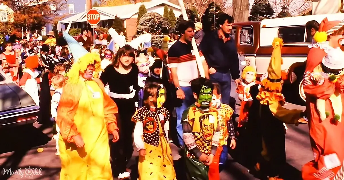 Children in costumes parade down a street with adults and cars in the background.