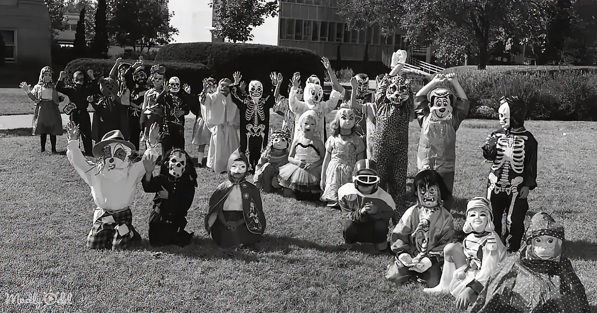 A group of people in various costumes, including skeletons and animals, pose for a photo on a grassy lawn.