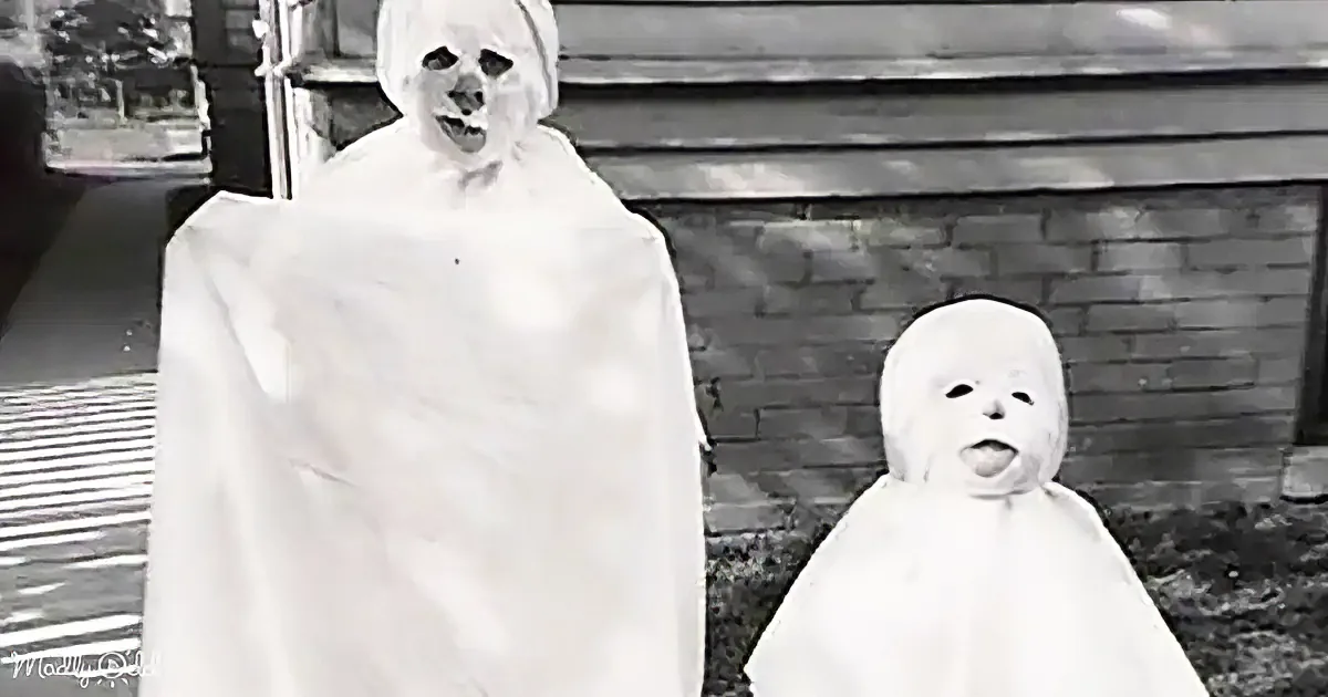 Two figures in ghost costumes stand side by side, one adult and one child, in front of a brick wall.