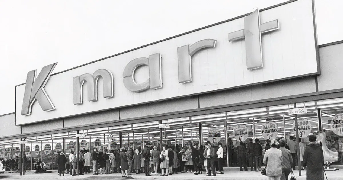A bustling Kmart storefront with a large crowd of people outside, some holding signs.