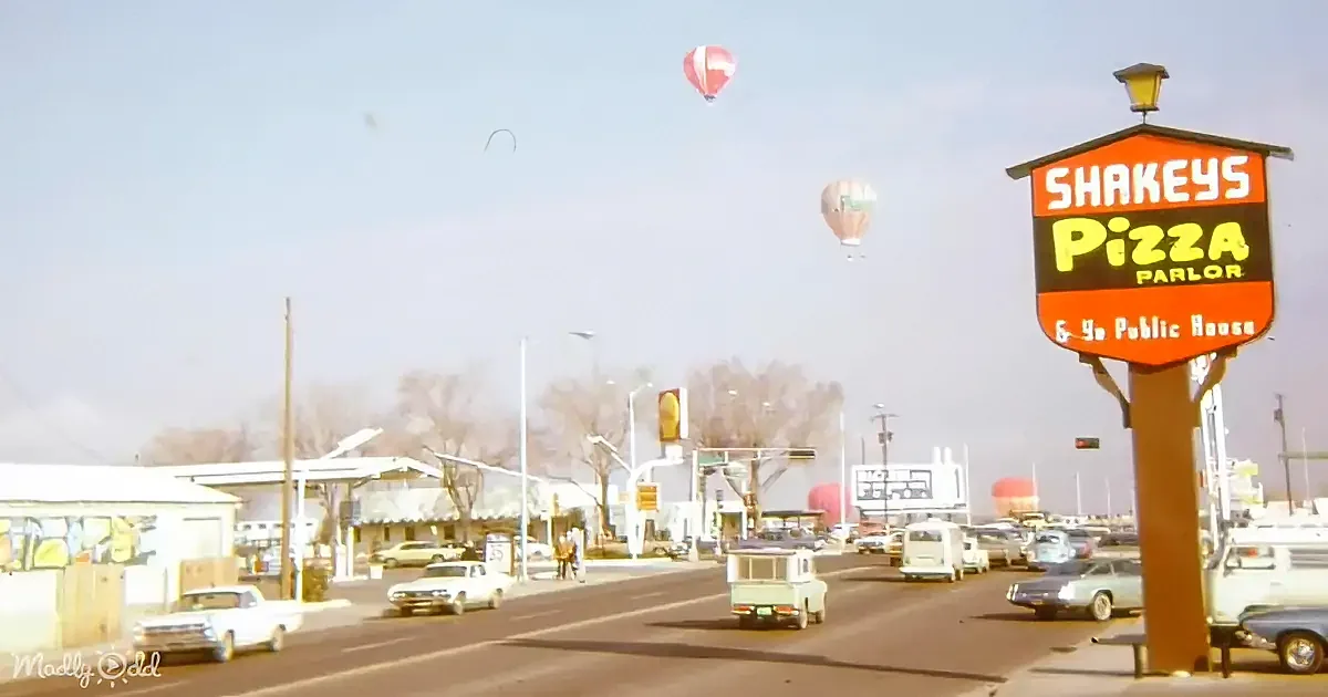Hot air balloons float above a Shakey's Pizza Parlor with vintage cars and a clear sky.
