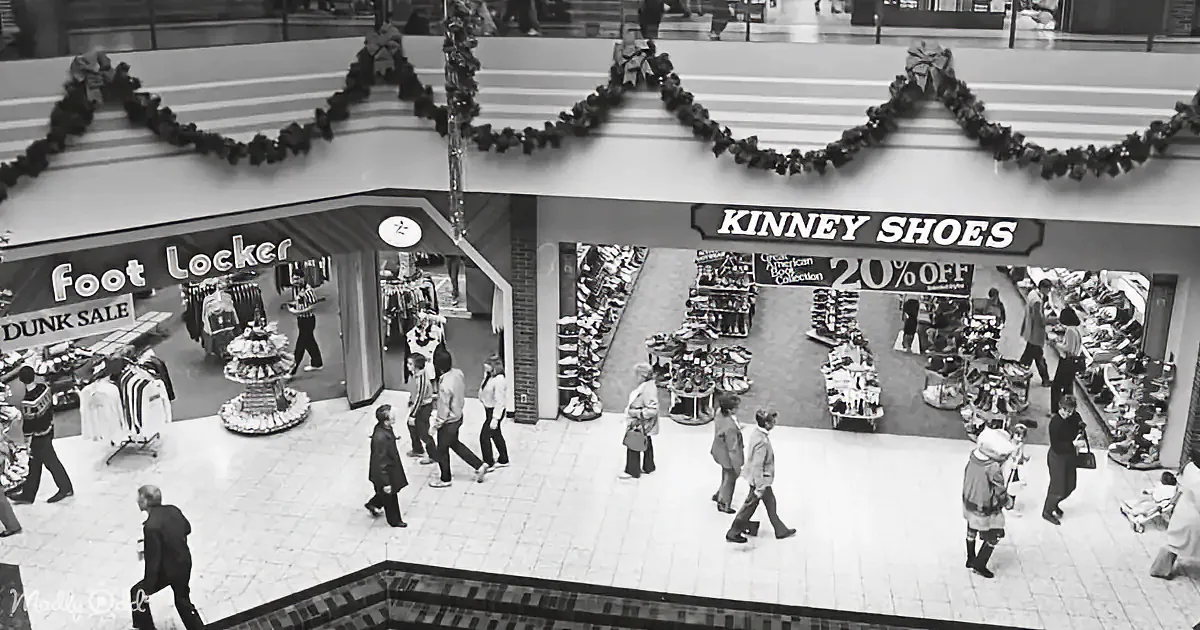 People walk past Kinney Shoes and Foot Locker in a bustling mall with festive decorations.