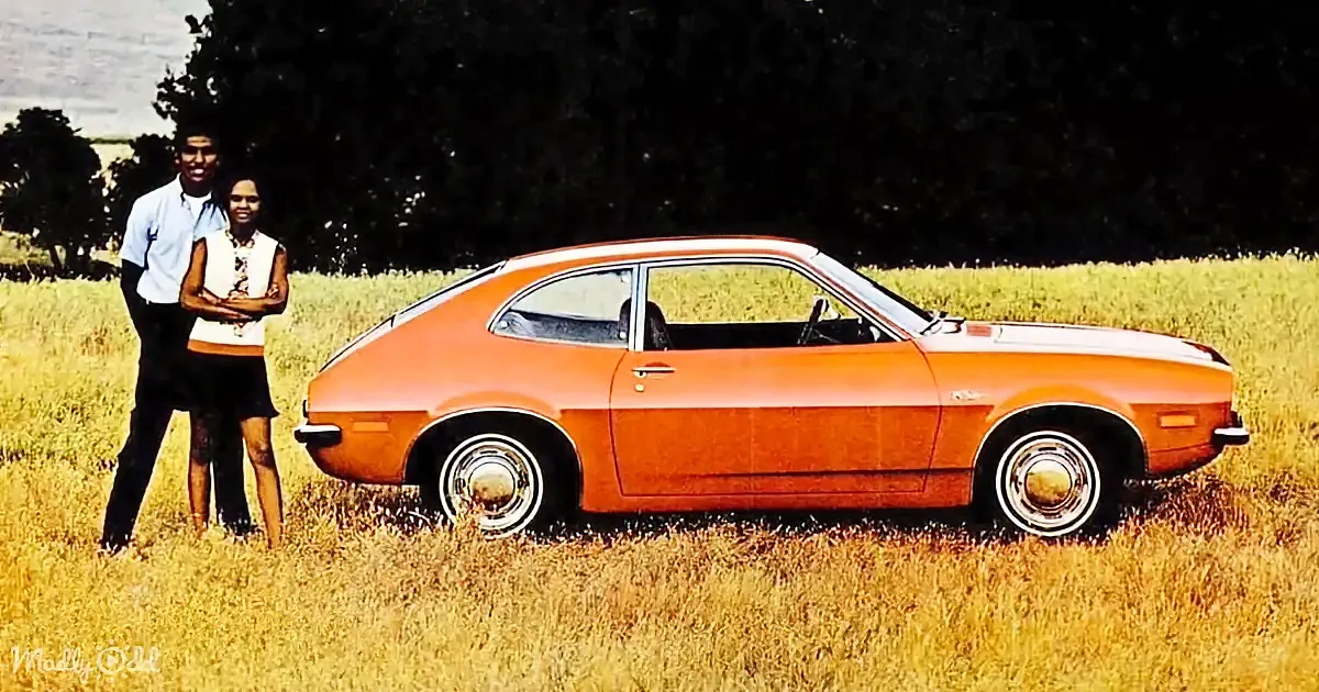 A couple stands beside an orange vintage car in a grassy field, surrounded by trees.