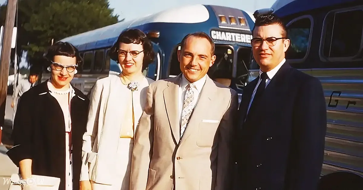 Four people in formal attire smile in front of a vintage charter bus on a sunny day.