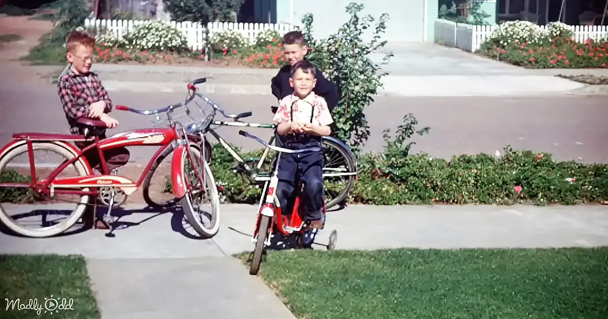 Two boys with bicycles, one on a red bike, the other on a smaller bike, in a suburban setting with a white picket fence...
