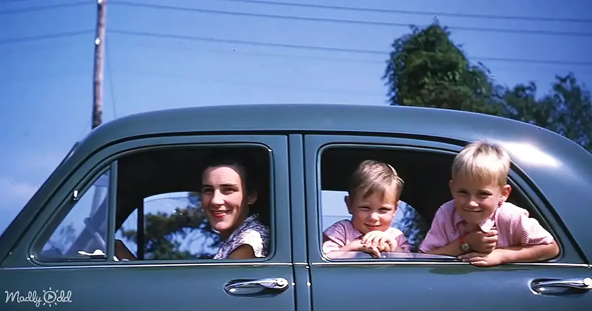 A woman and two children smile from the windows of a vintage car under a clear blue sky.