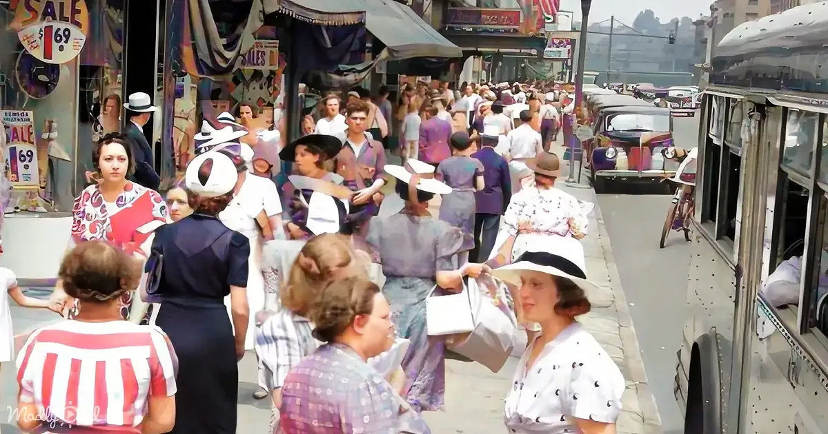 A bustling street scene with people in vintage clothing, hats, and a bus, surrounded by shops and cars.