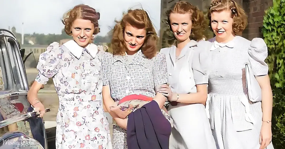 Four women in vintage dresses smile and pose outdoors near a car.
