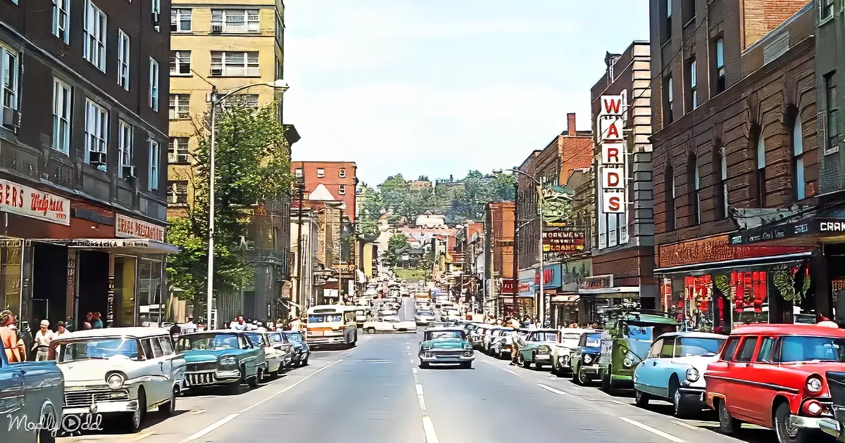 Vintage cars line a bustling street with old brick buildings and colorful storefronts under a bright sky.
