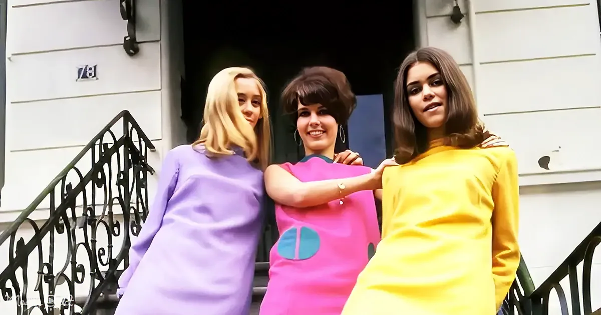 Three women in colorful dresses stand on a porch, smiling and posing together.