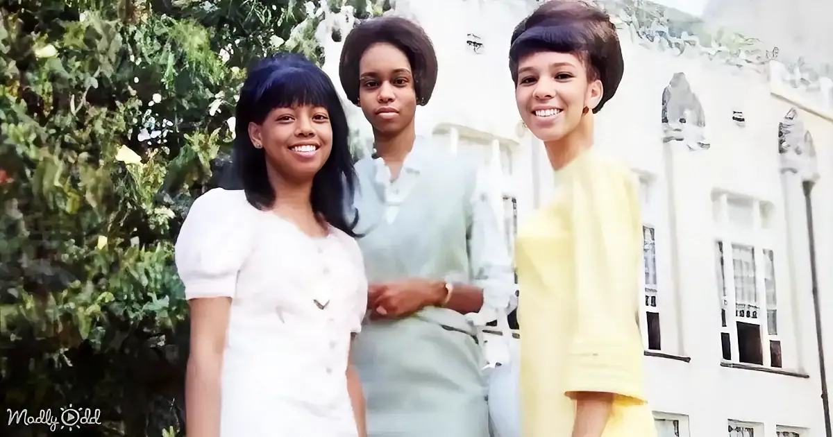 Three women in vintage attire smile in front of a white building with greenery.