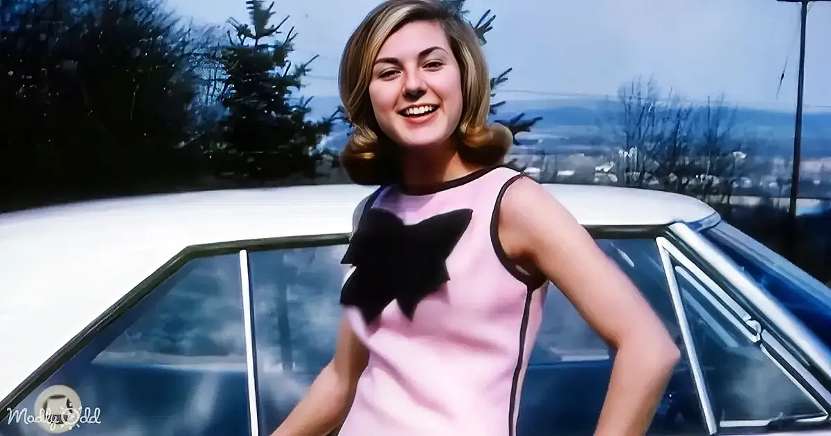 A woman in a pink dress with a black bow stands confidently in front of a car, smiling.