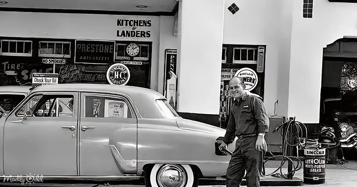 A man stands beside a vintage car in a garage filled with automotive memorabilia.