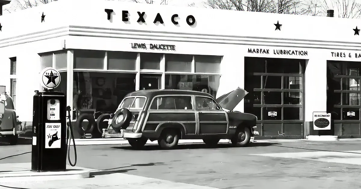A vintage car with its hood open is parked at a Texaco gas station, surrounded by classic service station elements.