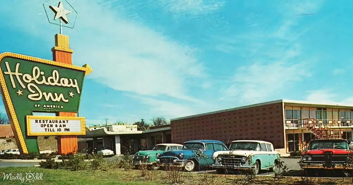 Holiday Inn sign with vintage cars parked in front of a mid-century motel under a blue sky.