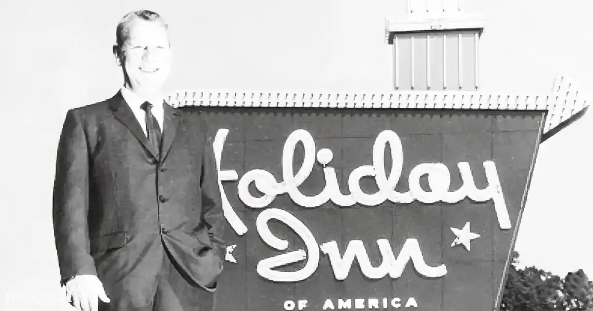 A man in a suit stands beside a sign reading "Holiday Inn" with a star and "of America" below.