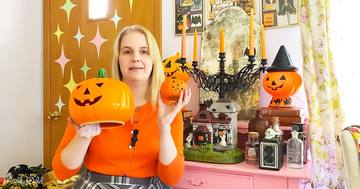 A person in an orange shirt holds two jack-o'-lanterns, surrounded by Halloween decorations and a pink cabinet.