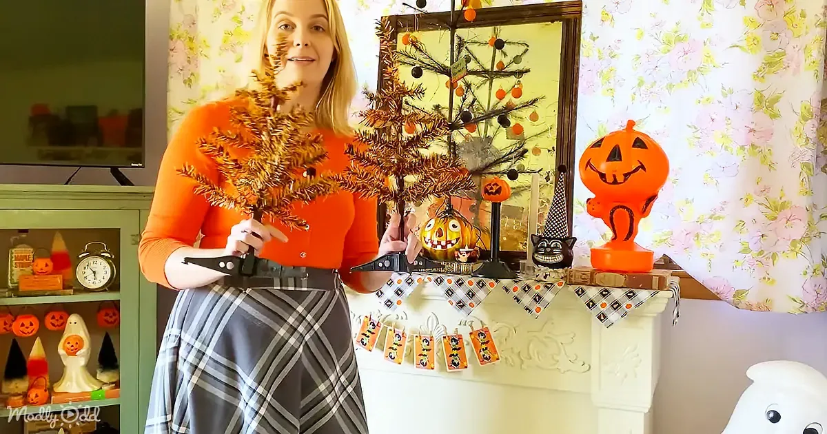 A woman in an orange top holds a small, tinsel tree, surrounded by Halloween decorations on a mantelpiece.
