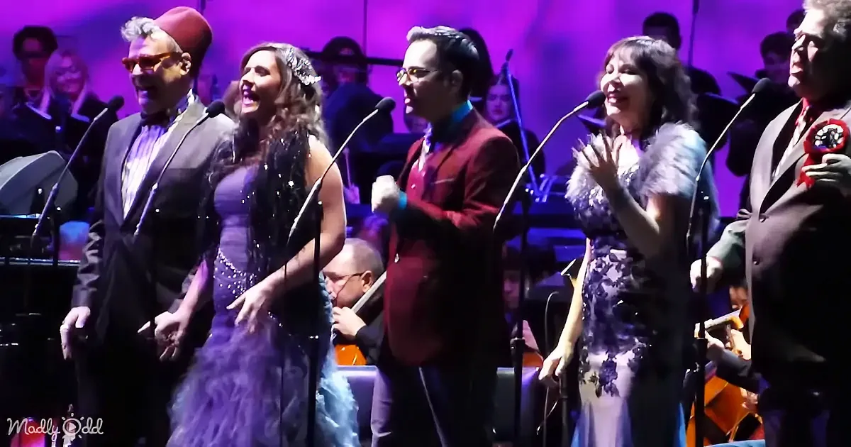 A group of performers in elegant attire sing on stage with a live orchestra in the background.