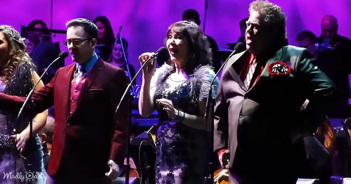 A woman in a fur-trimmed dress sings into a microphone, flanked by two men in suits, with a band in the background.