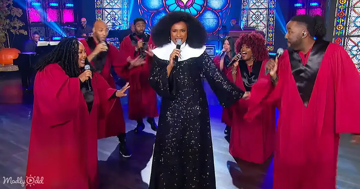 A woman in a black sequined dress sings into a microphone, surrounded by a choir in red robes.