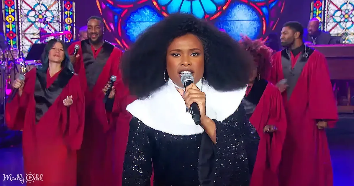 A woman with a large afro sings into a microphone, surrounded by a choir in red robes.