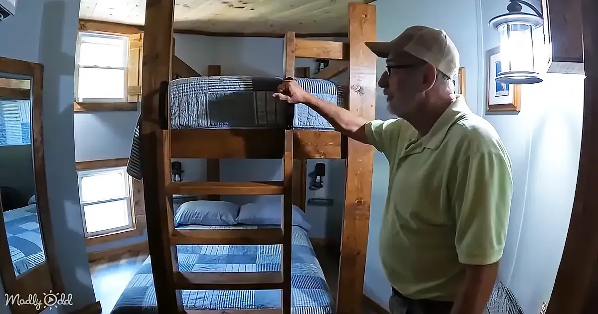 A man in a green shirt and cap points at a bunk bed in a cozy, well-lit room with wooden beams and windows.