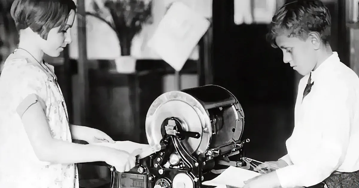 A woman operates an old-fashioned printing press while a man observes, both focused on the task.