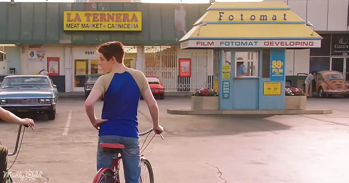A boy on a bike stands in front of a "La Ternera Meat Market" and a "Fotomat" film developing stand.