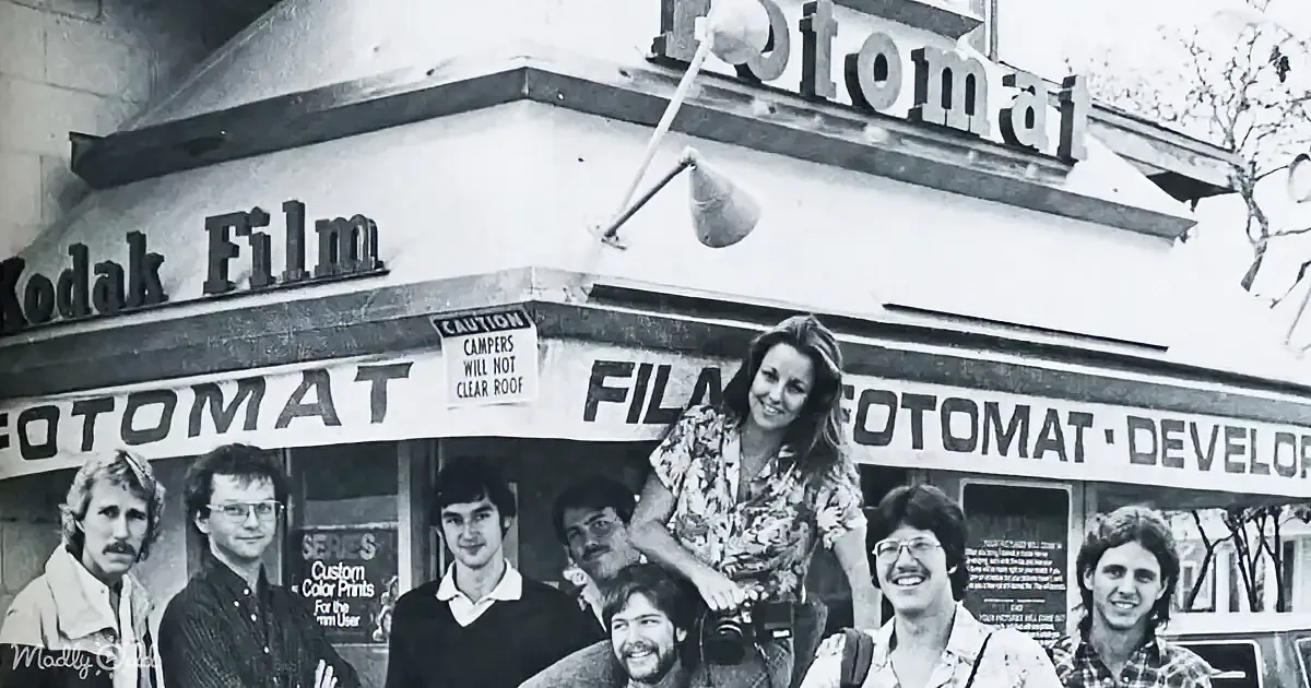 A group of people stands in front of a Fotomat film development shop, smiling and posing for a photo.