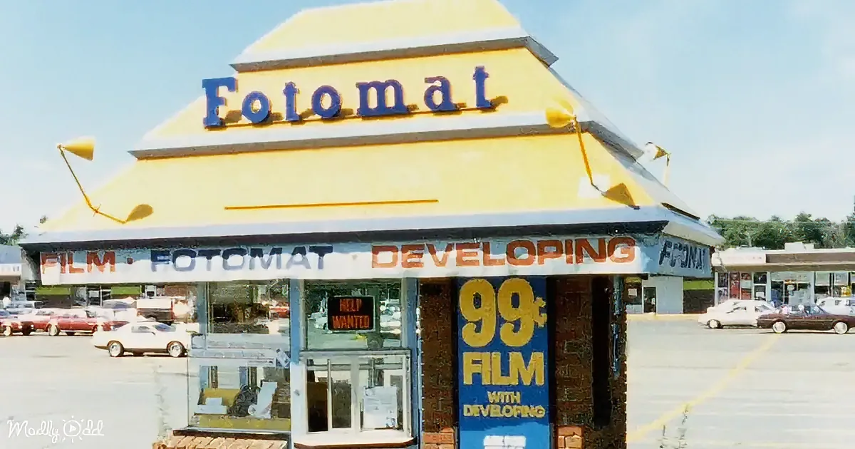 A vintage Fotomat store with a yellow sign and "99¢ Film with Developing" sign, parked cars, and a clear sky.