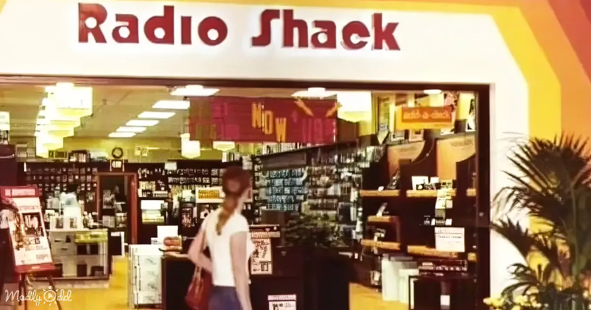 A woman walks into a Radio Shack store with shelves of electronics and a "Now Hiring" sign.