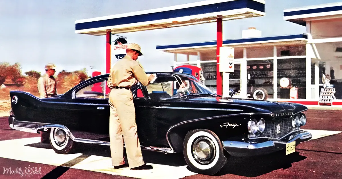A man in uniform stands beside a classic black car at a gas station, checking the vehicle.