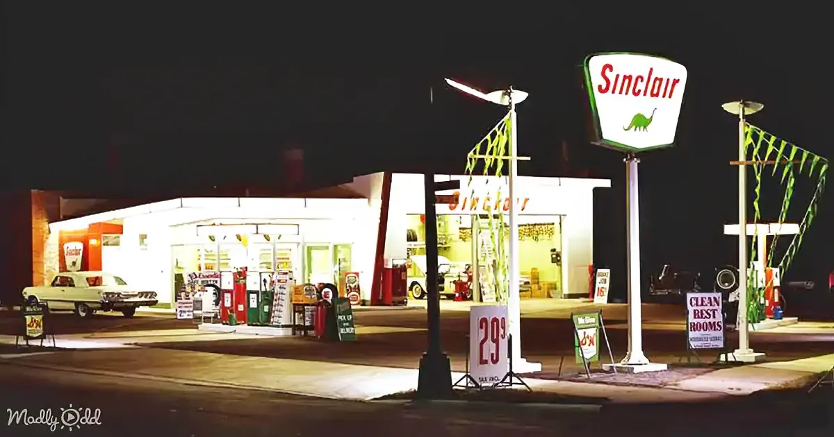 A vintage Sinclair gas station at night with illuminated pumps, a dinosaur sign, and a classic car parked outside.